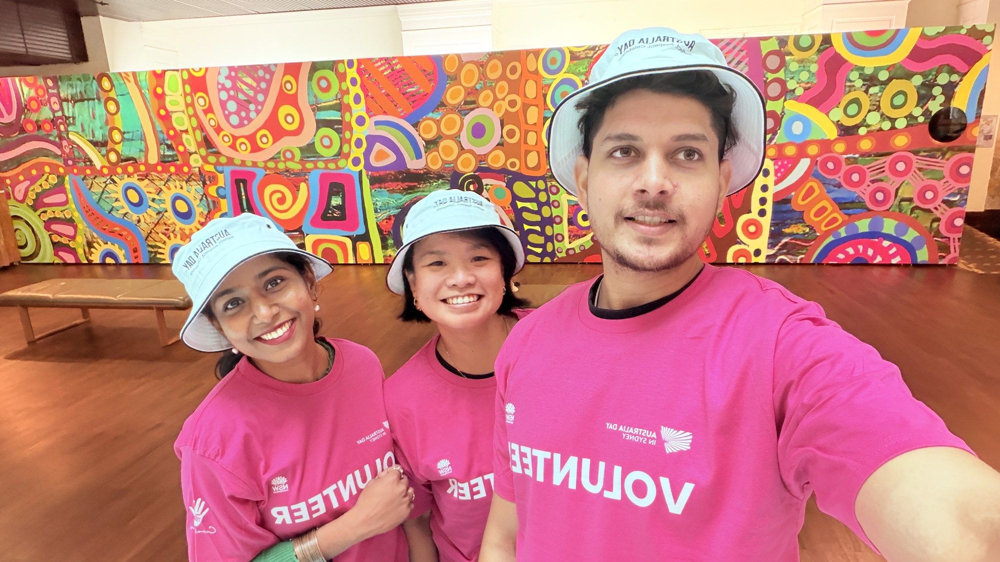 Manish and two volunteers in pink shirts and bucket hats in front of a bright Aboriginal art mural.