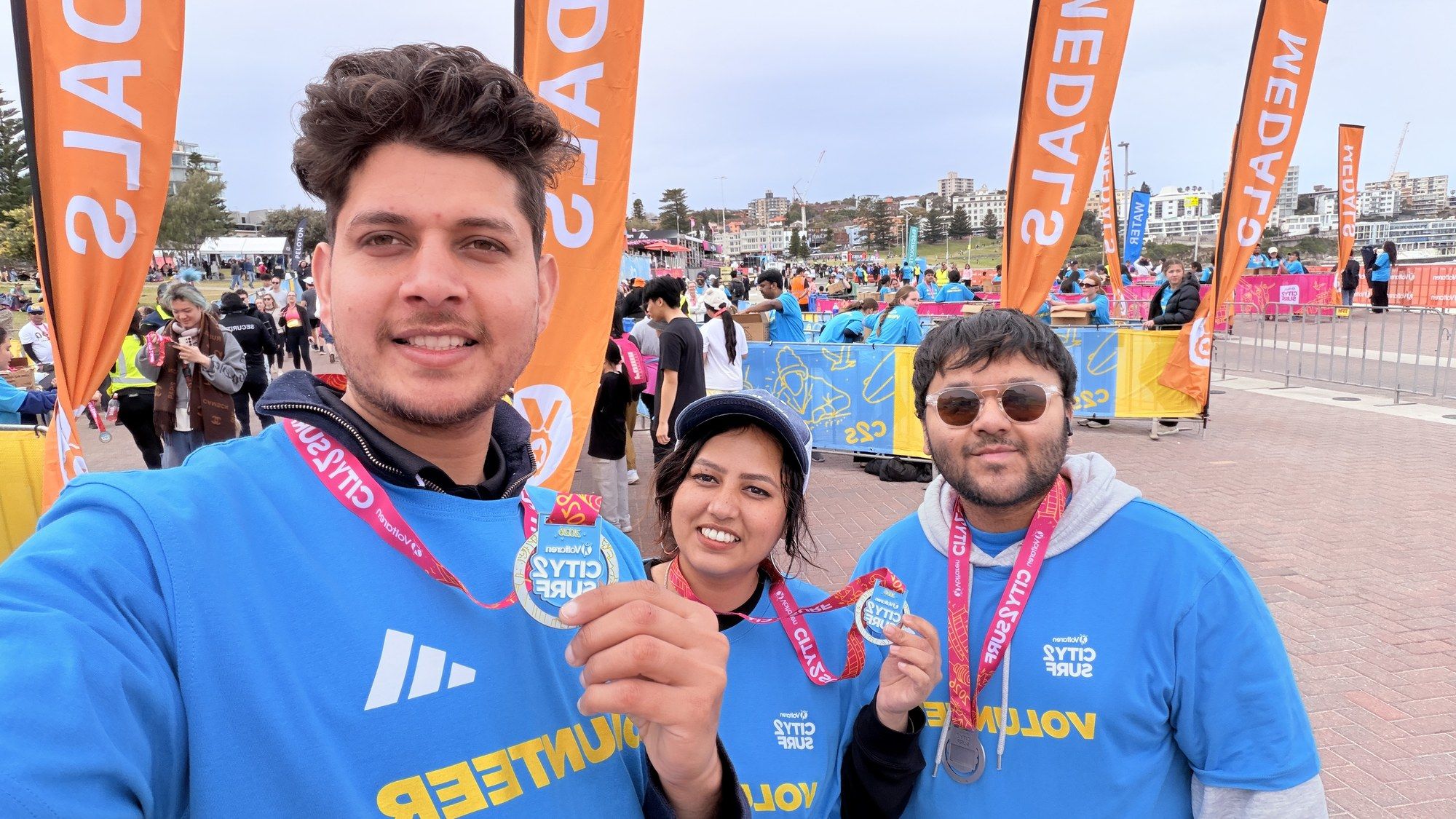 Manish and two volunteers holding up their City2Surf 2026 medals at Bondi.
