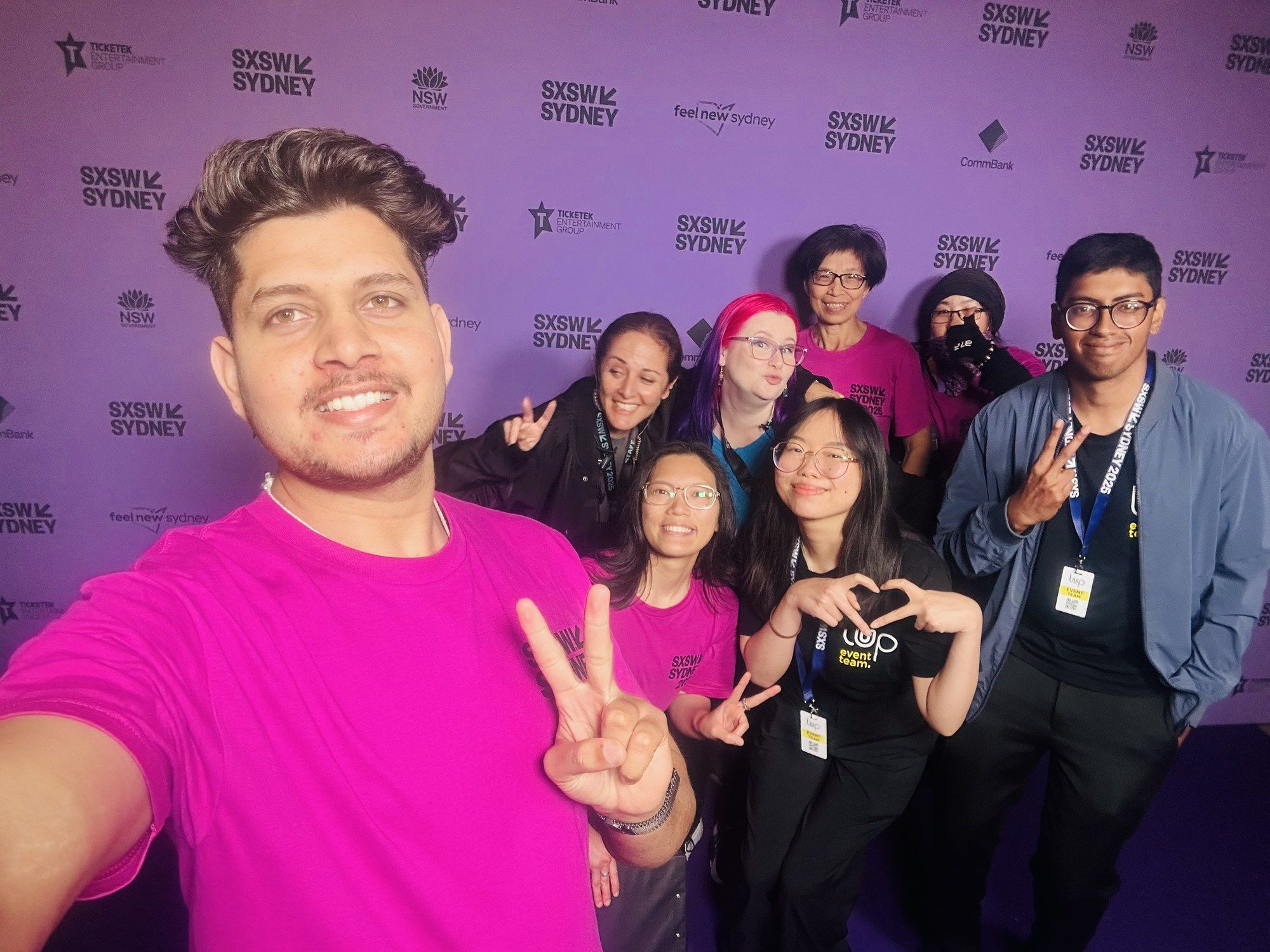 Manish taking a selfie with a group of SXSW Sydney volunteers in front of a sponsor wall.
