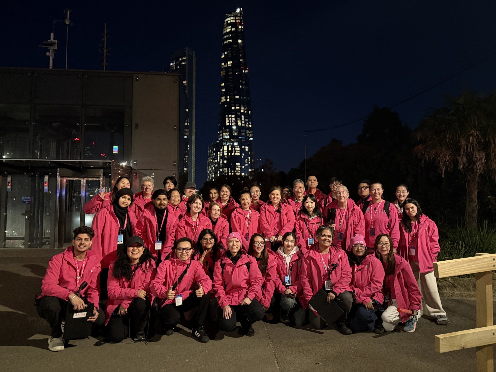 The whole Vivid Sydney volunteer crew in pink jackets, posed in rows at night with city towers behind.